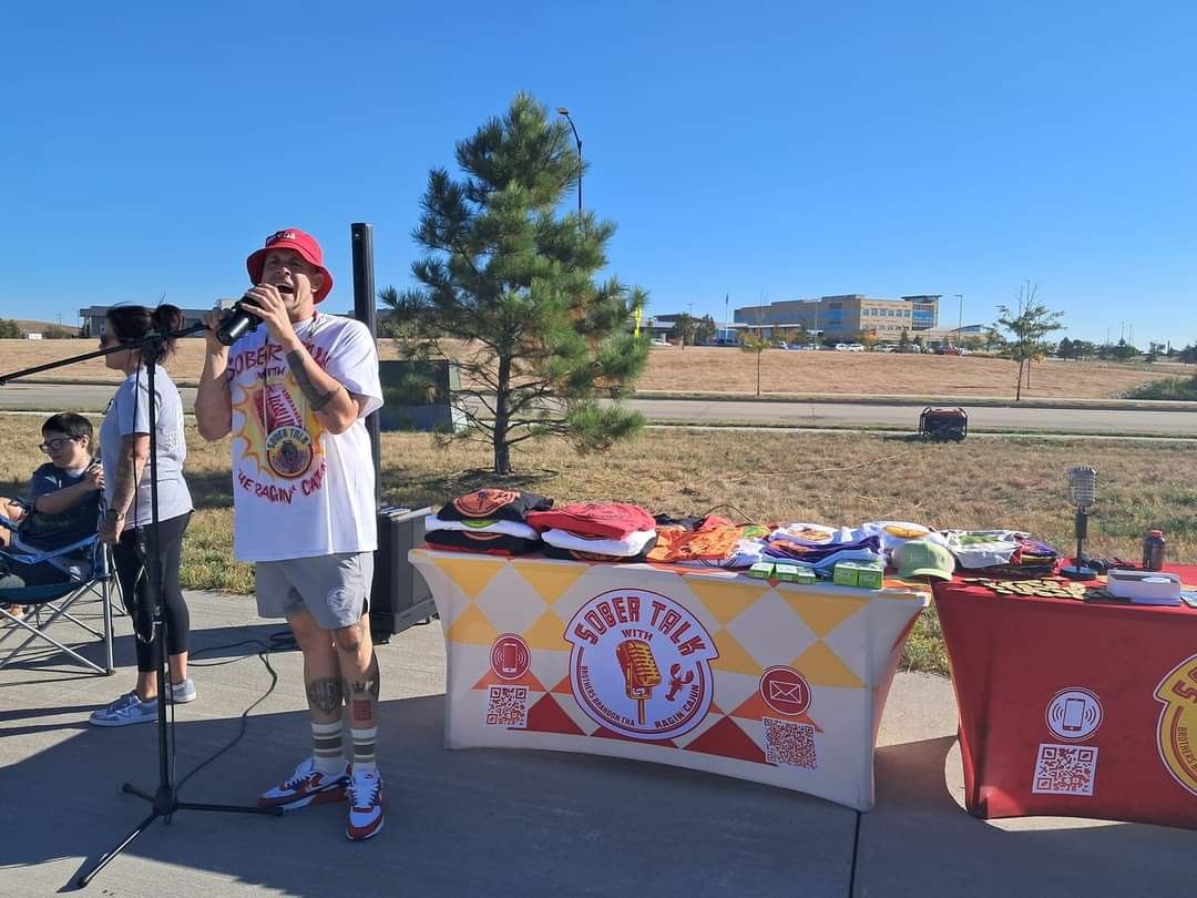 Brandon on the mic beside the SoberTalk table at an outdoor event